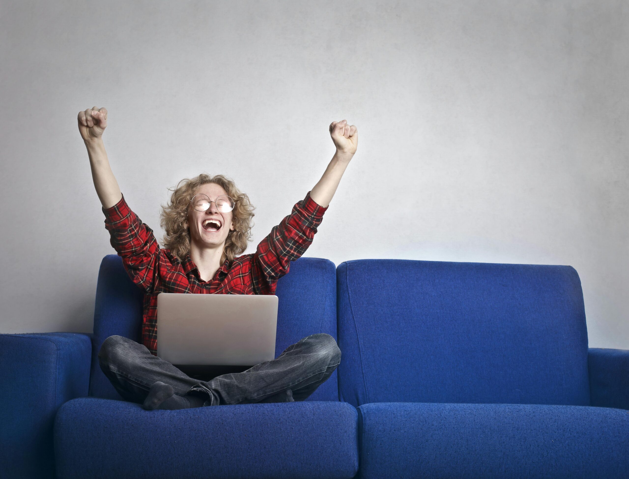 Foto Der Aufgeregten Person Mit Den Händen Oben, Die Auf Einem Blauen Sofa Sitzen, Während Sie Einen Laptop Benutzen